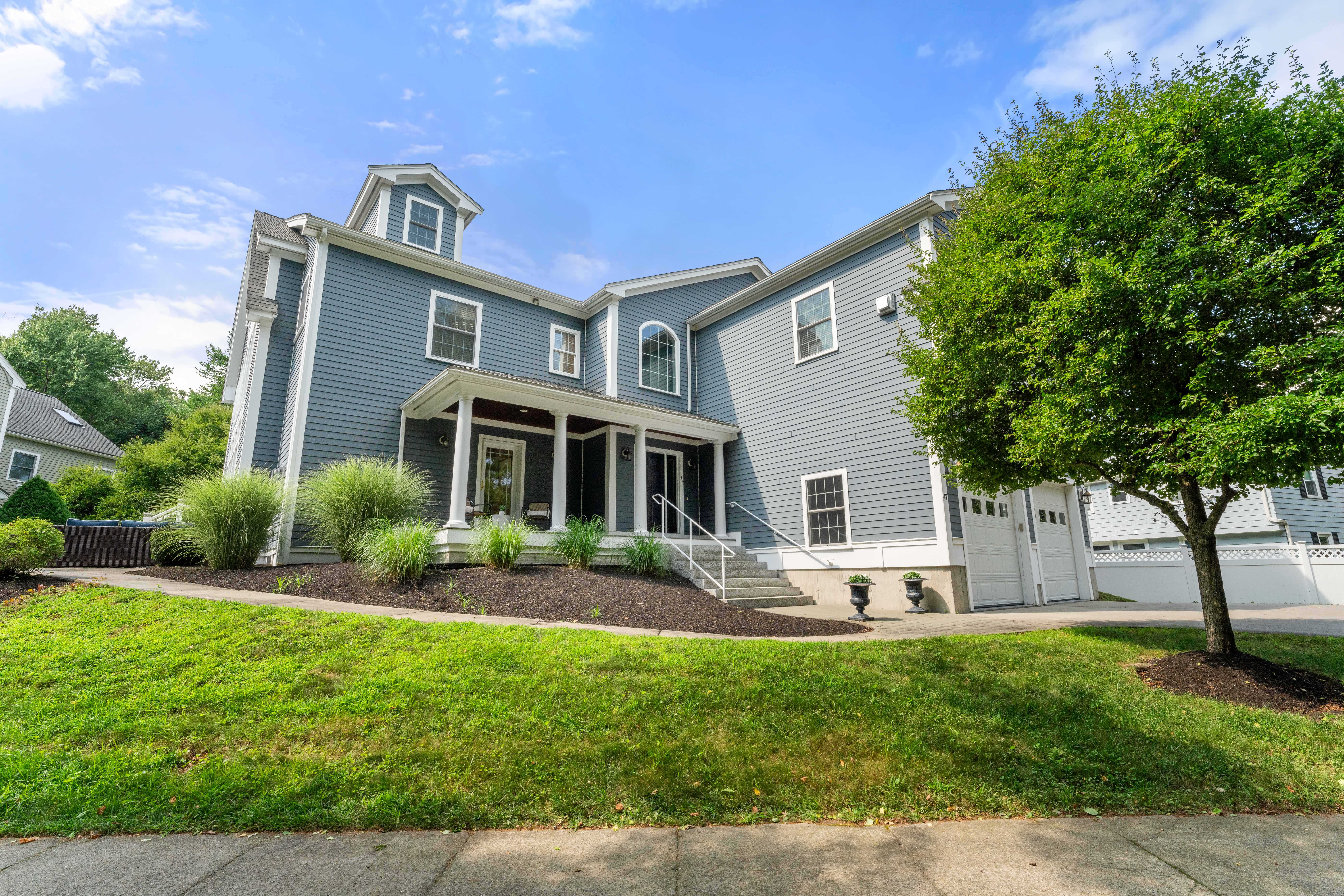 Exterior of a spacious blue-gray Colonial home in Needham, MA, featuring white trim, a welcoming covered front porch, two-car garage, manicured landscaping, and a mature shade tree.
