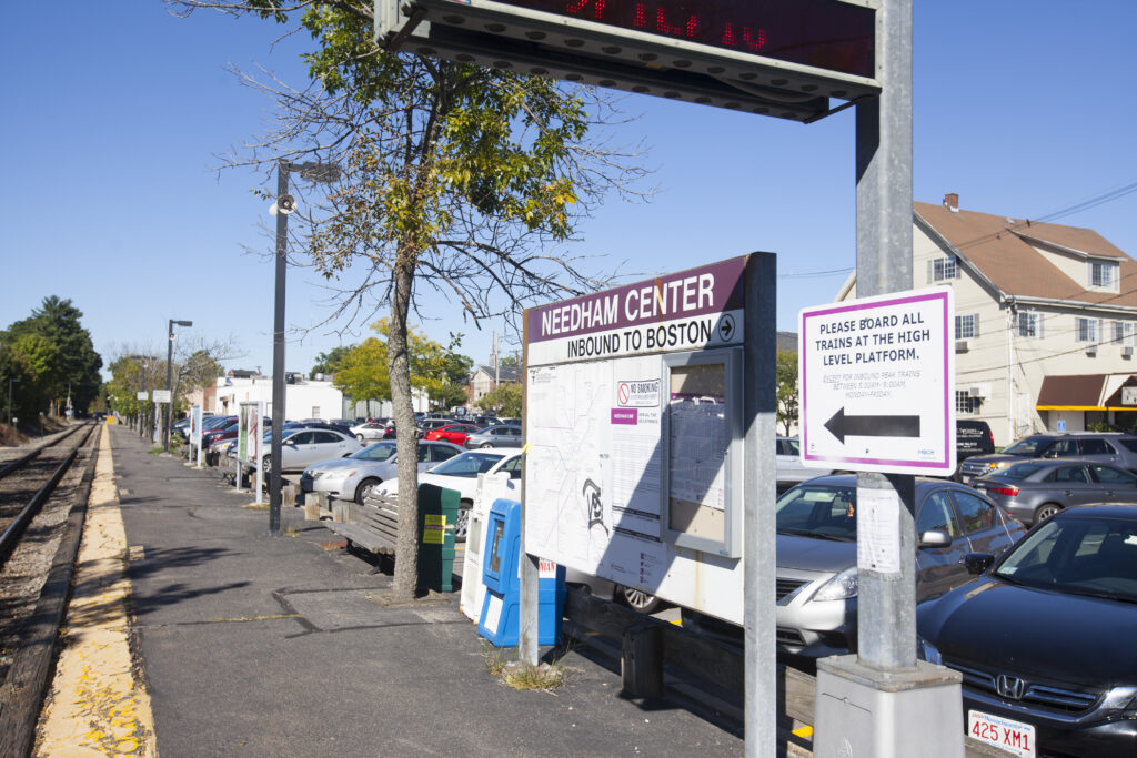 Needham Center MBTA commuter rail station platform in Needham, Massachusetts, with station signage, parked cars, and tracks leading toward Boston.